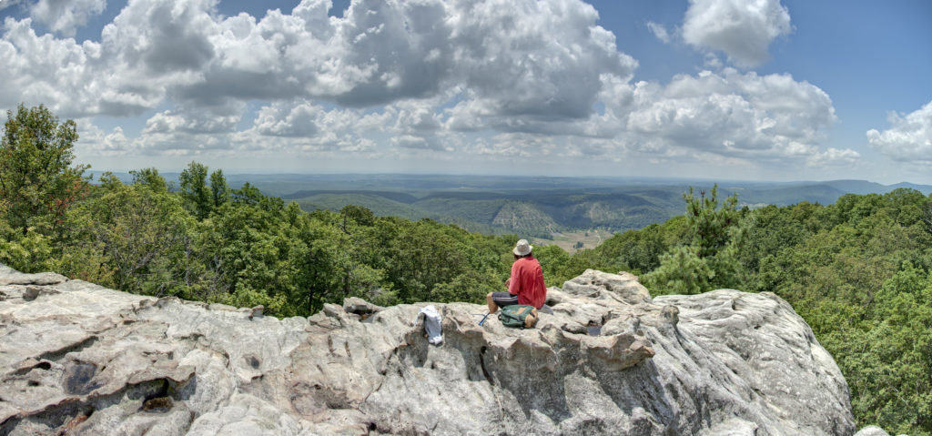 Hiker sitting atop a rocky bluff overlooking green treetops and a blue sky with puffy clouds