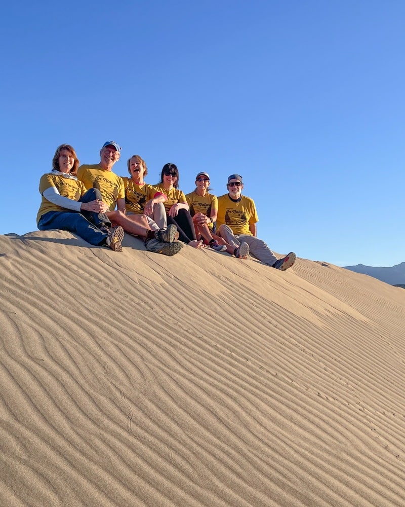 A six-person group smiling and sitting atop sandy hills in Death Valley, California, and surrounded by blue skies